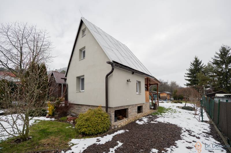 Záhradná chatka Handlová - Chata v Handlovej s trávnikom a zasneženou záhradou, obklopená stromami a kríkmi.
