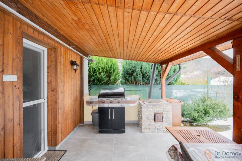 Záhradná chatka Handlová - Drevené obloženie a gril na terase chaty v Handlovej, vedľa zelených kríkov.