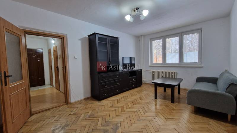 AstonReal:  predaj 2-izbový byt 54 m2, Poprad - Západ, Komenského - Obývacia izba dvojizbového bytu s pohovkou, tmavým nábytkom a podlahou s dreveným dekorom.