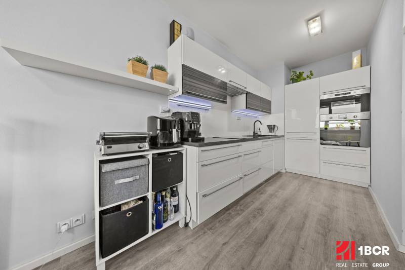 KOMPLETNE ZARIADENÝ 2-IZB. BYT,BALKÓN + PIVNICA,BUDATÍNSKA – PETRŽALKA - Kuchyňa v 2-izbovom byte s bielymi skrinkami a podlahou s dreveným dekorom.