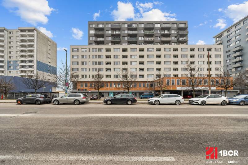 KOMPLETNE ZARIADENÝ 2-IZB. BYT,BALKÓN + PIVNICA,BUDATÍNSKA – PETRŽALKA - Budova na ulici Budatinská v Bratislave - Petržalke s autami zaparkovanými pred ňou.