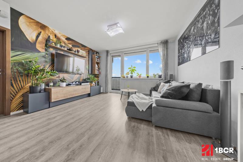 KOMPLETNE ZARIADENÝ 2-IZB. BYT,BALKÓN + PIVNICA,BUDATÍNSKA – PETRŽALKA - Obývacia izba s pohodlným gaučom, tapetou s rastlinným motívom a podlahou s dreveným dekorom.