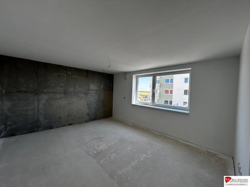 Predaj 3-izbového bytu len 15 minút od Bratislavy  - Prázdna izba v 3-izbovom byte s betónovou stenou a veľkým oknom.