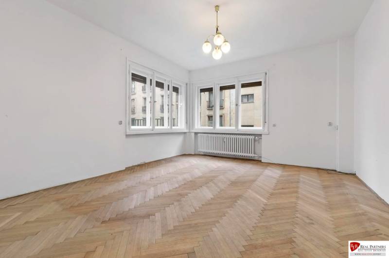 Predaj 4-izbový byt, 102m2, terasa 50m2, Bratislava-centrum, Gorkého - Izba v 4-izbovom byte so svetlom lustrom, podlahou s dreveným dekorom a veľkými oknami.