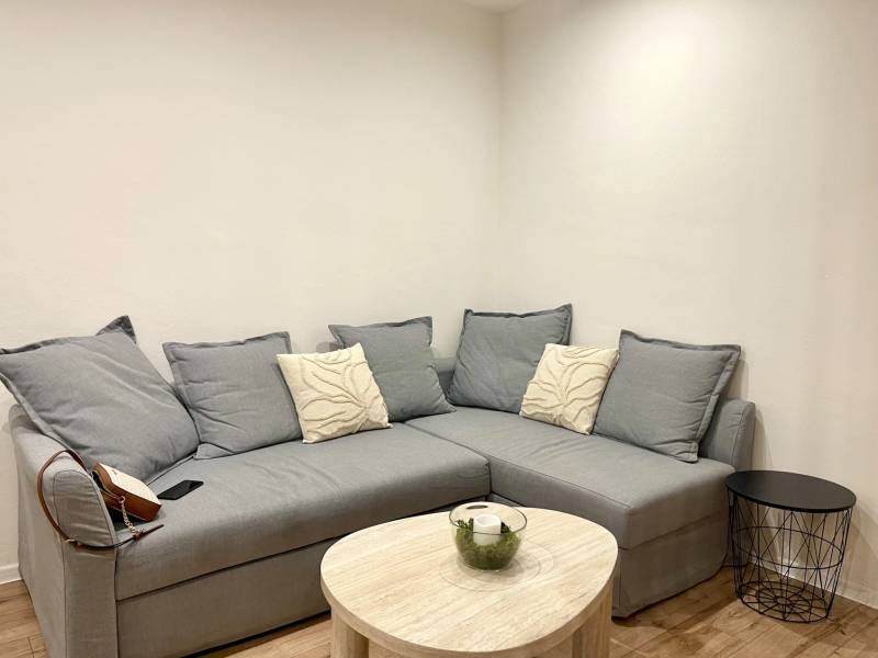 | Predaj 1-izbového bytu – Račianska, Bratislava - Sivá pohovka s vankúšmi a stolom v 1-izbovom byte s podlahou s dreveným dekorom.