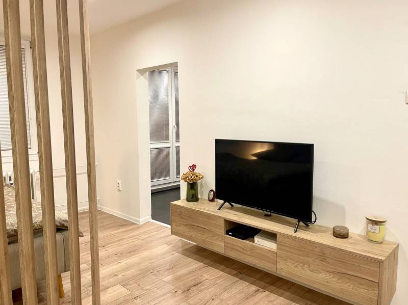 | Predaj 1-izbového bytu – Račianska, Bratislava - Izba s TV, podlahou s dreveným dekorom a stolíkom v 1-izbovom byte.