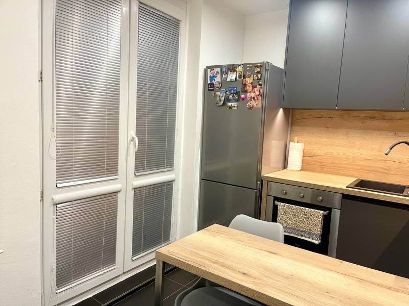 | Predaj 1-izbového bytu – Račianska, Bratislava - Kuchyňa v 1-izbovom byte s drevodekorom, sivý nábytok, veľké okno, chladnička.