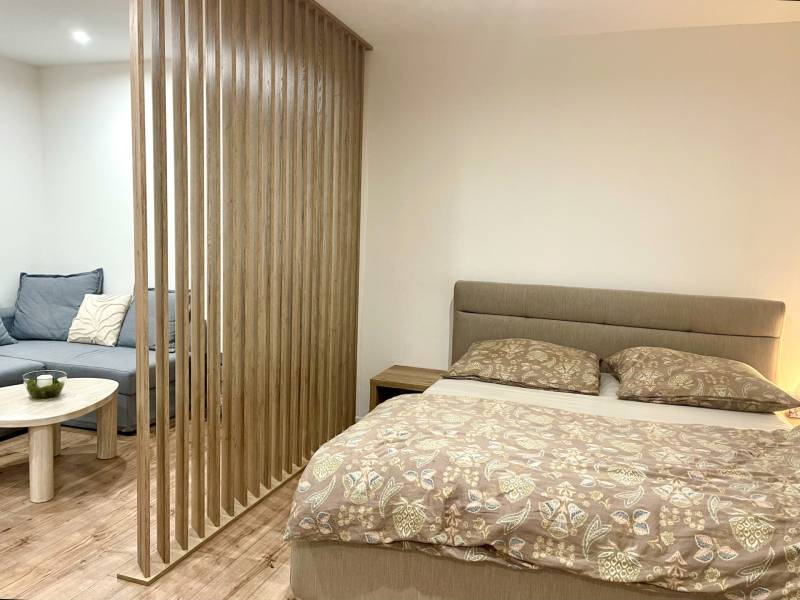 | Predaj 1-izbového bytu – Račianska, Bratislava - Jednoizbový byt s posteľou, sedačkou, dreveným panelom a podlahou s dreveným dekorom.