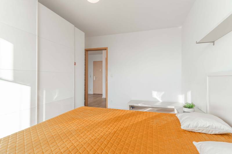 I na predaj 2i byt v komplexe Tri Veže na Bajkalskej - Spálňa v 2-izbovom byte s oranžovou prikrývkou a veľkou bielou skriňou.