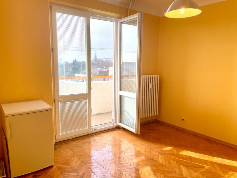 Predáj 1i čiastočne zrekonštruovaný byt s loggiou - Izba s podlahou s dreveným dekorom, radiátorom, oknom a dverami vedúcimi na balkón.