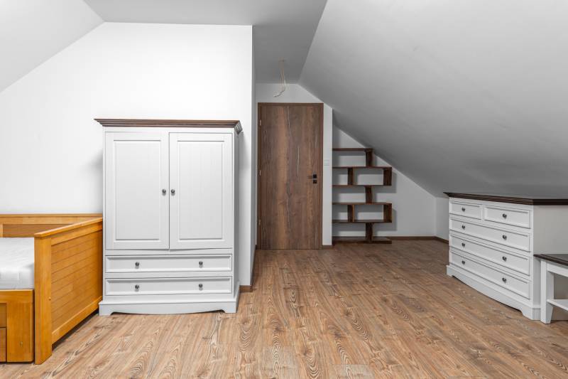 I Predaj štýlového tehlového domu v Rajke pri Bratislave - Biela skriňa, posteľ, zásuvky a poličky v podkroví rodinného domu s podlahou s dreveným dekorom.