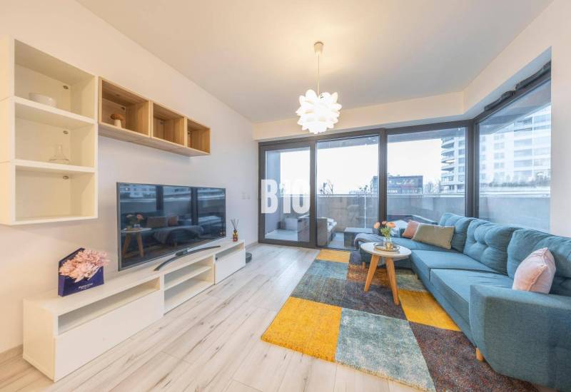 TEHELNÉ POLE | 1-izbový byt s priestrannou terasou - Obývacia izba v 1-izbovom byte s veľkou pohovkou, televízorom a podlahou s dreveným dekorom.