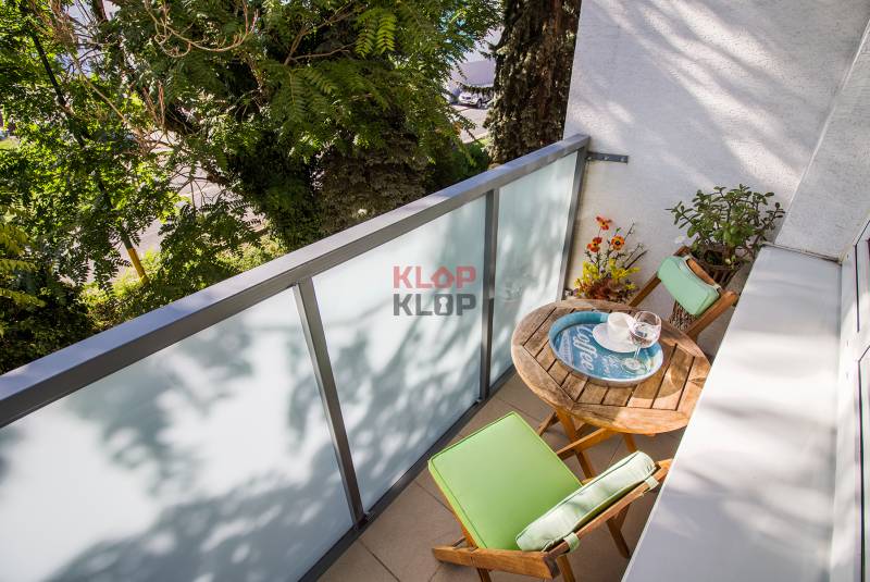 Pozrite si PRÍJEMNÝ 2i byt so ZARIADENÍM, LODŽIOU a KLIMATIZÁCIOU - Balkón 2-izbového bytu s dreveným stolom a stoličkami na ulici Mierová, Bratislava.