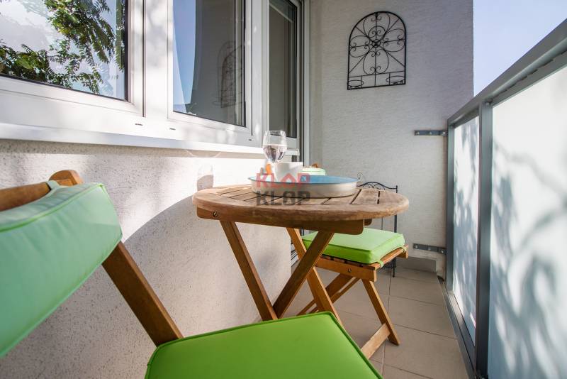 Pozrite si PRÍJEMNÝ 2i byt so ZARIADENÍM, LODŽIOU a KLIMATIZÁCIOU - Balkón v 2-izbovom byte s dreveným stolom, stoličkami a dekoračnou kovovou mriežkou.