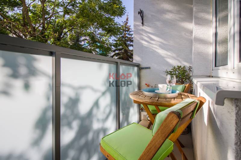 Pozrite si PRÍJEMNÝ 2i byt so ZARIADENÍM, LODŽIOU a KLIMATIZÁCIOU - Balkón 2-izbového bytu s dreveným nábytkom, zelenými vankúšmi a výhľadom na zeleň.