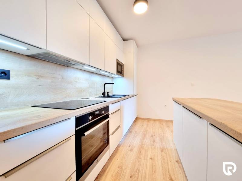 Pekný a moderný 3-izbový byt I Arboria Trnava - Kuchyňa v 3-izbovom byte s podlahou s dreveným dekorom, bielymi skrinkami a vstavanými spotrebičmi.