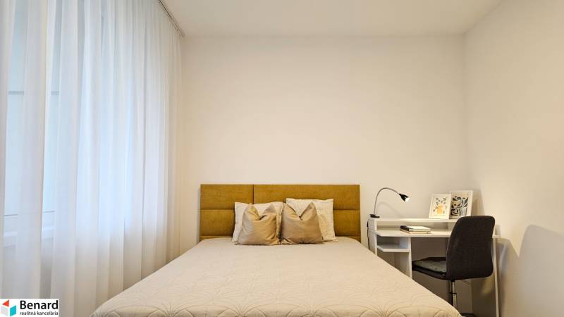 Moderný unikát - 1,5- izb. byt s 2 loggiami na prenájom (Nad jazerom) - Spálňa v 1-izbovom byte so svetlou posteľou, záclonami a bielym stolom s lampou.