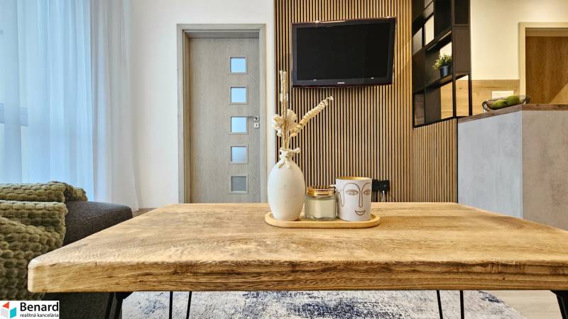 Moderný unikát - 1,5- izb. byt s 2 loggiami na prenájom (Nad jazerom) - Obývacia časť 1-izbového bytu s dreveným stolom a dekoráciami na pozadí s televízorom.