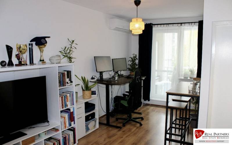 Prenájom 2-izb bytu Dlhé diely - Obývacia izba s policou plnou kníh a trofejí, pracovným stolom a kobercovou podlahou.