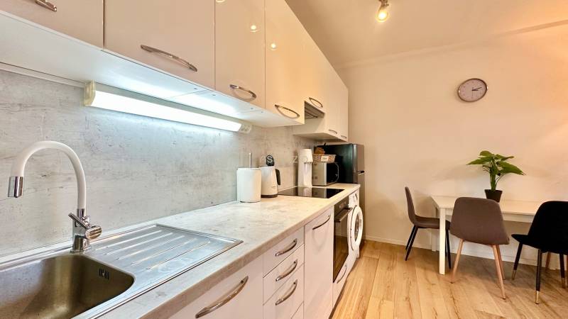 – PRENAJOM – 2 IZBOVÝ BYT 52 m² – OSTREDKOVA – RUŽINOV  - Kuchyňa v 2-izbovom byte s podlahou s dreveným dekorom, jedálenským stolom a spotrebičmi.