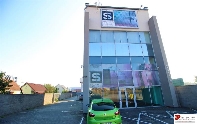 Predaj modernej administratívnej budovy v Bratislave, okres II - Budova so sklenenou fasádou, áutom v popredí a parkoviskom na ulici.