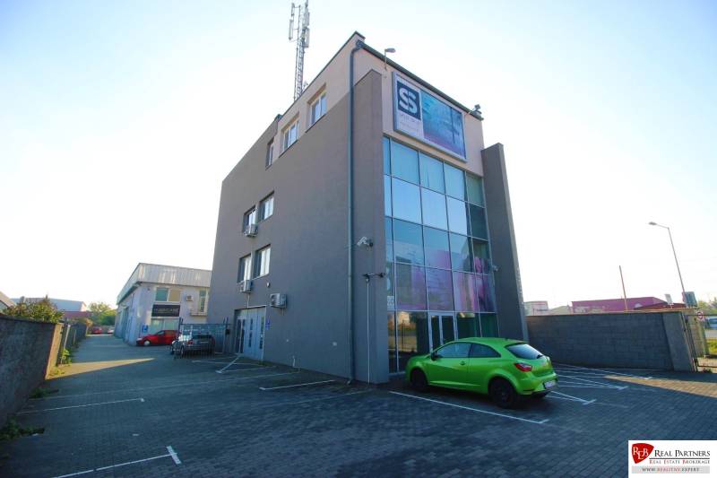 Predaj modernej administratívnej budovy v Bratislave, okres II - Budova so sklenou fasádou, parkoviskom a zelenej auto. Nachádza sa v priemyselnej oblasti.