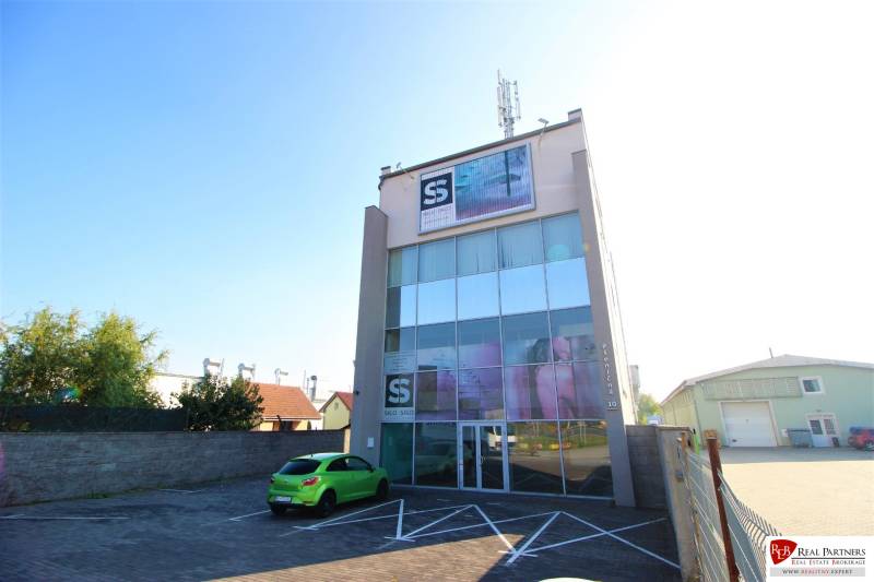 Predaj modernej administratívnej budovy v Bratislave, okres II - Budova s presklenou fasádou a anténou, zaparkované auto, slnečný deň na parkovisku.