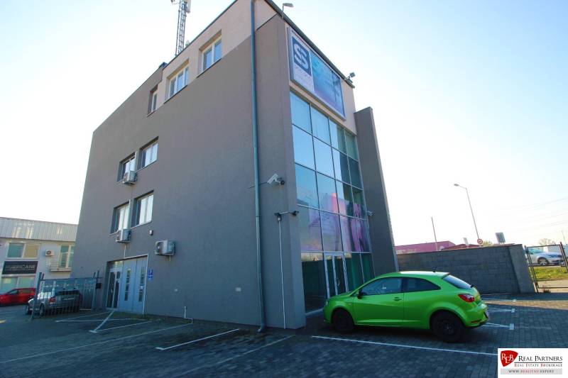 Predaj modernej administratívnej budovy v Bratislave, okres II - Budova s presklenou fasádou, zeleným autom a parkoviskom v exterieri.