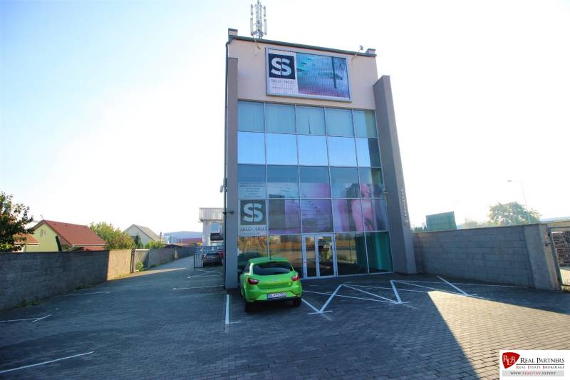 Predaj modernej administratívnej budovy v Bratislave, okres II - Štvorposchodová budova so sklenenou fasádou a parkoviskom pred vchodom.