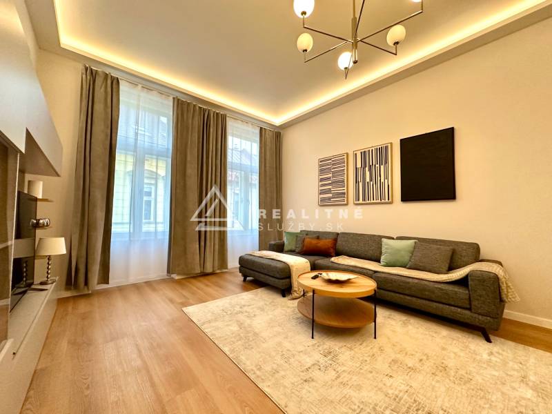 Prenájom priestranného 2-izbového bytu v absolútnom centre Košíc - Obývačka v 2-izbovom byte s podlahou s dreveným dekorom, šedou sedačkou a dekoratívnymi obrazmi.