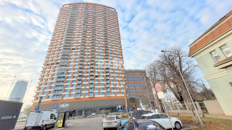 PRENÁJOM - VNÚTORNÉ PARKOVACIE STÁTIE, KLINGERKA, Súkennícka ulica - Veža v Bratislave - Ružinov na Súkenníckej ulici, obklopená autami a parkoviskom.