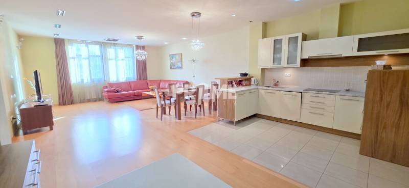 VEĽKOMETRÁŽNY 3i byt, 3 x NEPRIECHODNÁ IZBA, KLÍMA, BALKÓN, Nám.SNP - Obývacia izba v 3-izbovom byte s podlahou s dreveným dekorom, kuchyňou a červenou sedačkou.