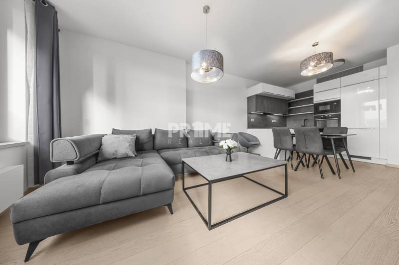 ŠTÝLOVÝ 2i byt,SKY PARK,VÝHĽAD,LOGGIA,PARKING,VEŽA II,Továrenská ul. - Obývačka v 2-izbovom byte s veľkou šedou sedačkou a kuchyňou, podlaha s dreveným dekorom.