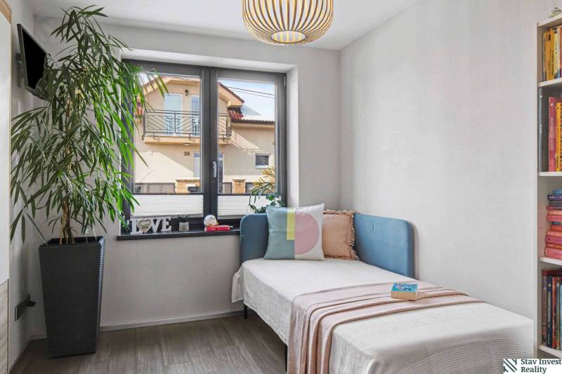 Ideálny dom pre rodinu, Blatné, okr. Senec - Izba v rodinnom dome s rastlinou, posteľou, vankúšmi a knižnicou. Oknom vidieť susedný dom.