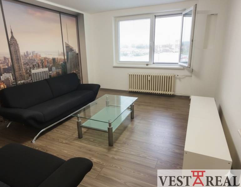 Na predaj: 2-izbový byt v centre mesta Galanta – časť Nová Doba - Obývačka v 2-izbovom byte s čiernym gaučom, skleneným stolom a podlahou s dreveným dekorom.