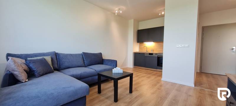 Zariadený 2-izb. byt v novostavbe v Ružinove - Ovocné sady - 2-izbový byt s modrou sedačkou, tmavou kuchyňou a podlahou s dreveným dekorom.