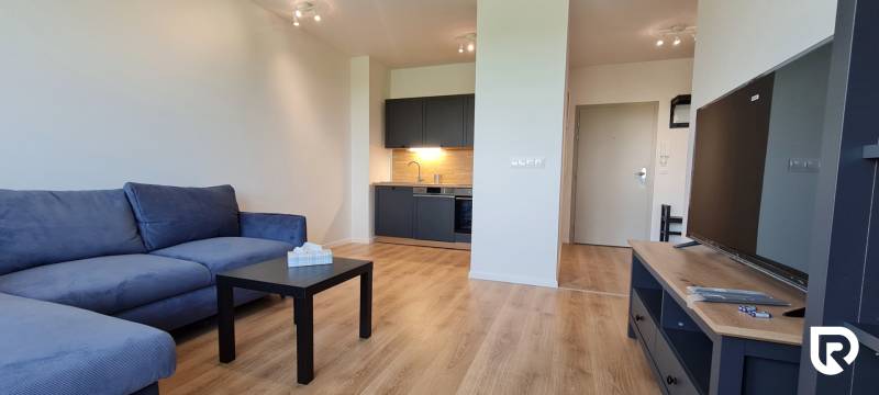 Zariadený 2-izb. byt v novostavbe v Ružinove - Ovocné sady - Obývačka v 2-izbovom byte s modrou sedačkou, kuchynským kútom a podlahou s dreveným dekorom.