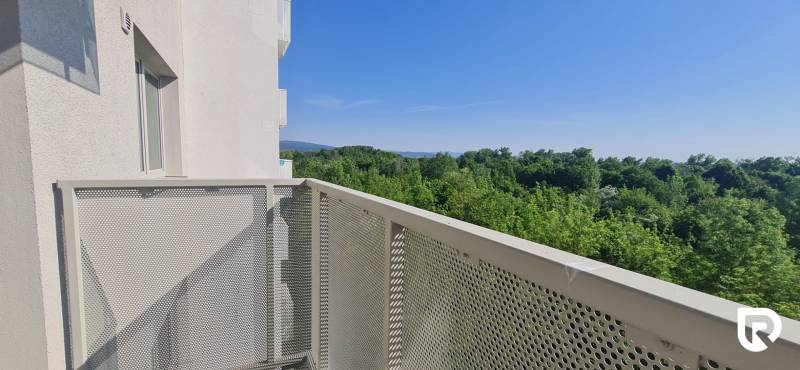 Zariadený 2-izb. byt v novostavbe v Ružinove - Ovocné sady - Balkón s výhľadom na les a kopce, budova v pozadí, jasná obloha.
