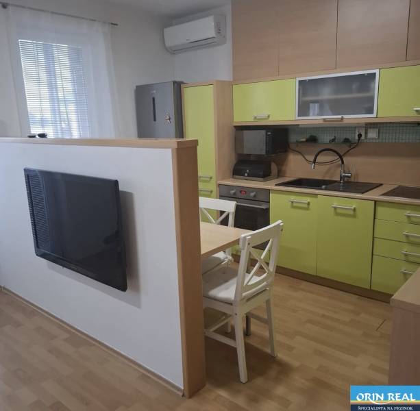 Na prenájom klimatizovaný 2 izbový byt v Moste pri Bratislave - Kuchyňa v 2-izbovom byte so zelenými skrinkami, televízorom a stolom s dvomi stoličkami.