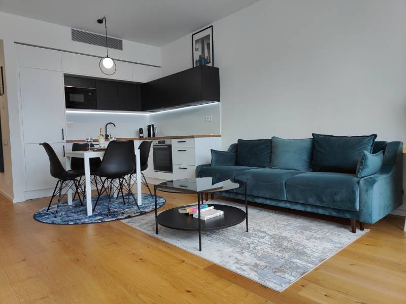 NUERA︱SKY PARK Moderný 2-izbový byt na 6. poschodí, veža 3 - Obývačka s kuchynským kútom, tyrkysovou sedačkou, dreveným dekorom a jedálenským stolom.