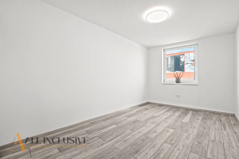 NA PRENÁJOM 3-IZB BYT S BALKÓNOM A ZÁHRADOU,DUN.STREDA - Prázdna izba, biela stena, podlaha s dreveným dekorom, okno s dekoratívnou rastlinou.