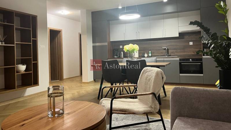 EXKLUZÍVNE-PREDAJ 3-IZBOVÝ BYT POPRAD, širšie centrum 80 m2 s gar.st. - Obývacia izba v 3-izbovom byte s kuchyňou, stolom a podlahou s dreveným dekorom.