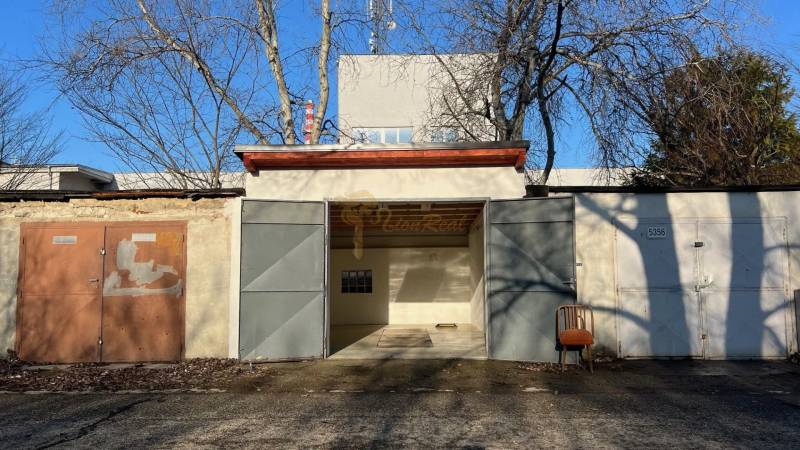 Zrekonštruovaná veľká garáž na ul. Agátova, Dúbravka s pozemkom - Garáž na Agátovej ulici v Bratislave - Dúbravke s plechovými dverami a stoličkou vonku.