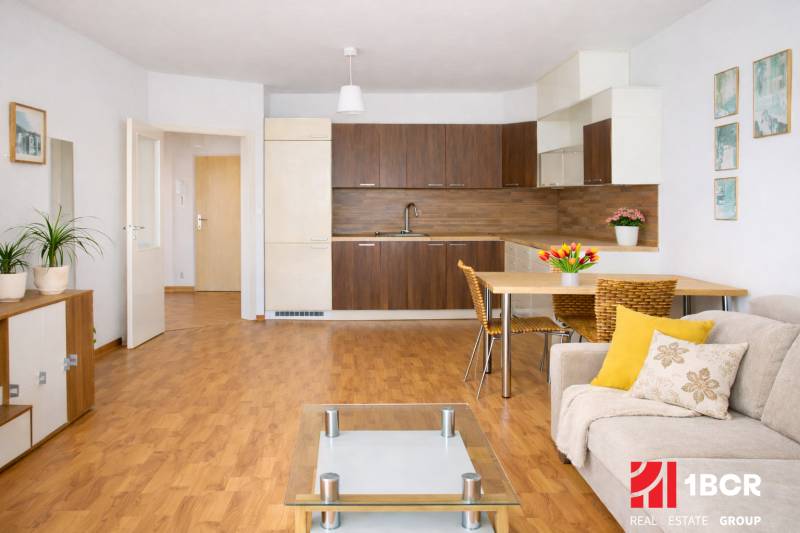 INVESTIČNÉ BÝVANIE V TOP LOKALITE: 2-IZBOVÝ BYT, JÉGÉHO UL, BRATISLAVA - Obývačka 2-izbového bytu s kuchyňou, podlahou s dreveným dekorom a pohodlným sedením.