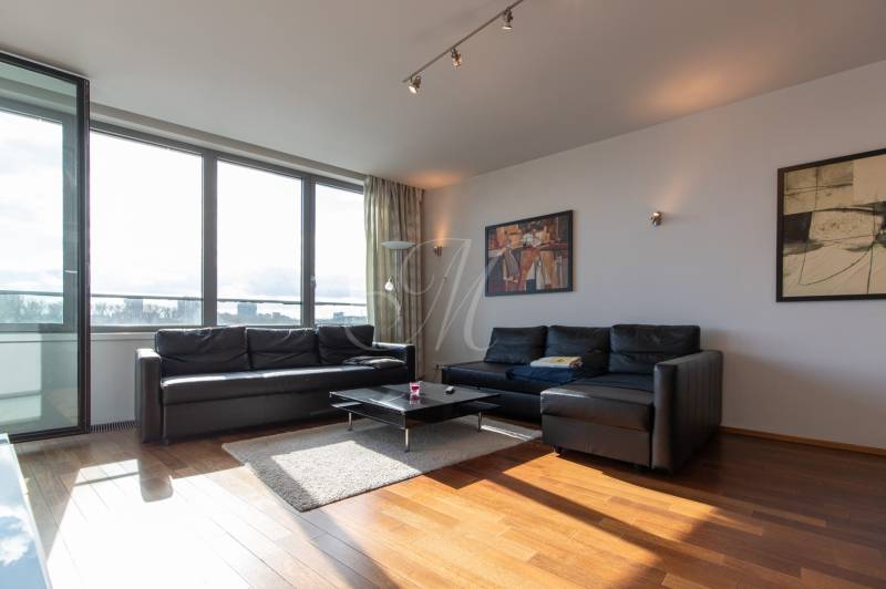 Moderný 2-izbový byt s balkónom, Eurovea - Obývačka v 2-izbovom byte s čiernou sedačkou, podlahou s dreveným dekorom a veľkými oknami.