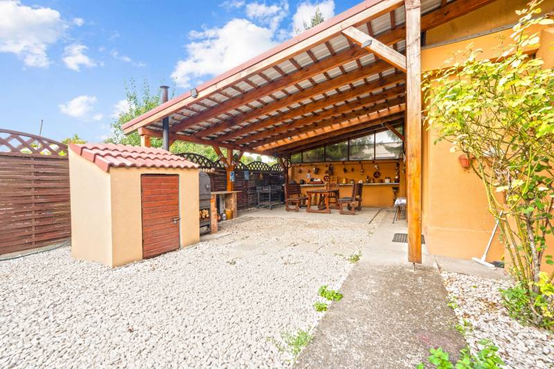 NA PREDAJ VÝNIMOČNÝ RODINNÝ DOM VO VEĽKOM MEDERI - Záhrada rodinného domu vo Veľkom Mederi s prístreškom a drevným dekorom na stene.