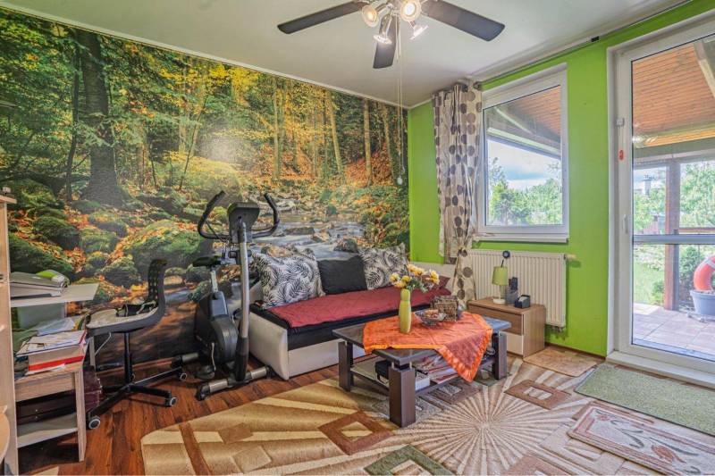 NA PREDAJ 3 IZBOVÝ RODINNÝ DOM, KĽÚČIAROVÉ KRAČANY - Obývacia izba v rodinnom dome s lesnou tapetou, stolíkom a podlahou s dreveným dekorom.