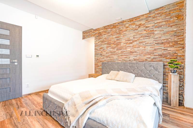 NA PREDAJ NADŠTANDARDNÁ 4-IZB. NOVOSTAVBA, DUN. KLÁTOV - Spálňa v rodinnom dome s kamennou stenou, elegantnou posteľou a dekoratívnymi doplnkami.