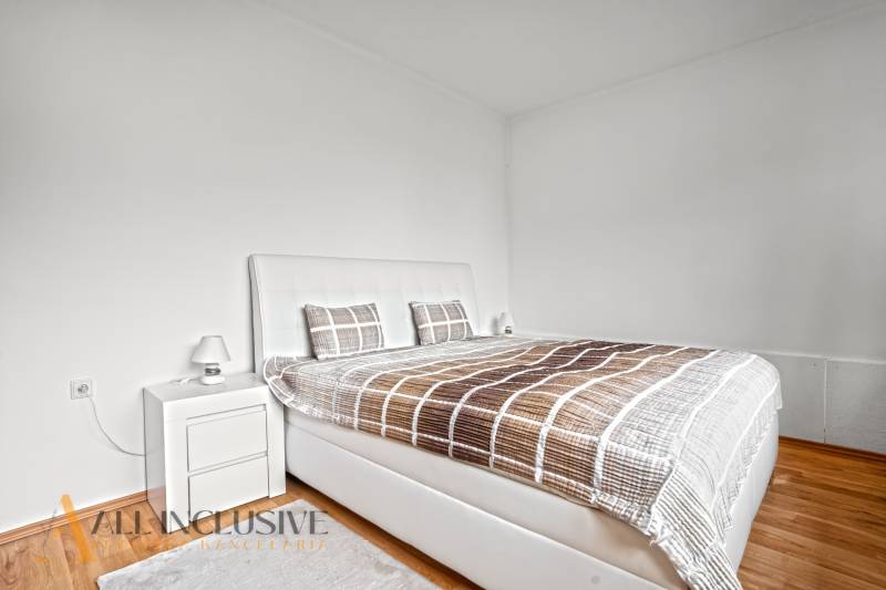 NA PREDAJ APARTMÁNOVÝ DOM VO VEĽKOM MEDERI - Svetlá spálňa s veľkou posteľou, podlahou s dreveným dekorom a nočným stolíkom.