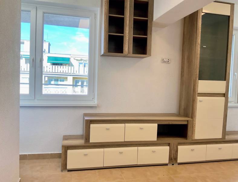 I Predaj Investičného 1-izbového bytu s veľkorysou loggiou  - Obývacia stena v 1-izbovom byte s podlahou s dreveným dekorom a výhľadom z okna.